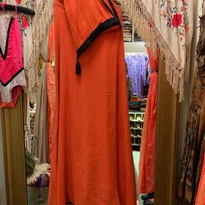 Puede incluir: Se muestra una prenda naranja con ribete negro. La prenda est&aacute; drapeada sobre una percha, mostrando su dise&ntilde;o fluido. El fondo incluye textiles decorativos y un espejo, lo que sugiere un entorno de boutique.