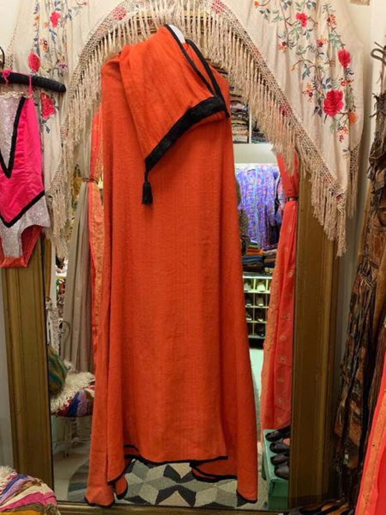 Puede incluir: Se muestra una prenda naranja con ribete negro. La prenda est&aacute; drapeada sobre una percha, mostrando su dise&ntilde;o fluido. El fondo incluye textiles decorativos y un espejo, lo que sugiere un entorno de boutique.