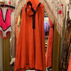 Puede incluir: Una capa con capucha naranja con ribete negro cuelga frente a un espejo. La capa tiene un dise&ntilde;o largo y fluido y una cremallera negra. Un leotardo rosa y blanco cuelga a la izquierda. El fondo incluye un tapiz floral y otras prendas.