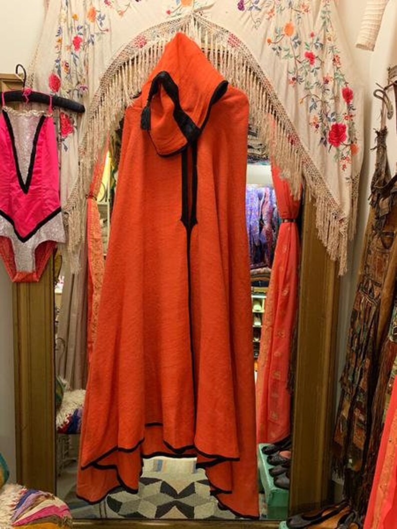 Puede incluir: Una capa con capucha naranja con ribete negro cuelga frente a un espejo. La capa tiene un dise&ntilde;o largo y fluido y una cremallera negra. Un leotardo rosa y blanco cuelga a la izquierda. El fondo incluye un tapiz floral y otras prendas.