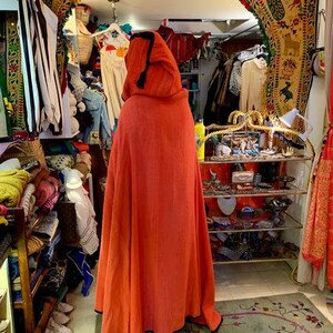 Puede incluir: Una capa con capucha naranja con ribete negro es el punto focal de esta imagen. La capa se exhibe en una tienda llena de ropa y accesorios. La tienda tiene un arco decorativo y una alfombra estampada. Otras prendas son visibles en el fondo.