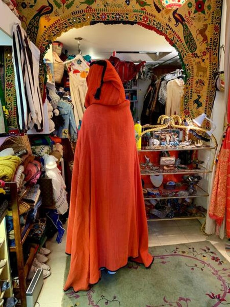 Puede incluir: Una capa con capucha naranja con ribete negro es el punto focal de esta imagen. La capa se exhibe en una tienda llena de ropa y accesorios. La tienda tiene un arco decorativo y una alfombra estampada. Otras prendas son visibles en el fondo.
