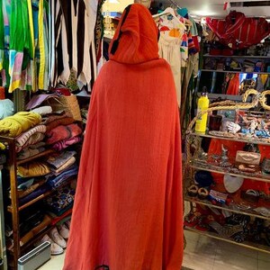 Puede incluir: Una capa con capucha de color naranja vibrante con ribete negro, exhibida en una tienda de ropa vintage. La capa est&aacute; hecha de una tela texturizada y cuelga suelta. Otras prendas son visibles en el fondo, incluyendo chaquetas y vestidos coloridos.
