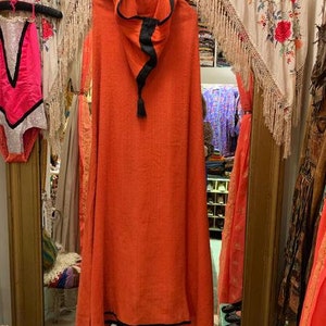 Puede incluir: Una capa con capucha naranja con ribete negro y borla. La capa est&aacute; colgada de una percha blanca en una tienda vintage. Un traje de ba&ntilde;o rosa con ribete negro cuelga a la izquierda.