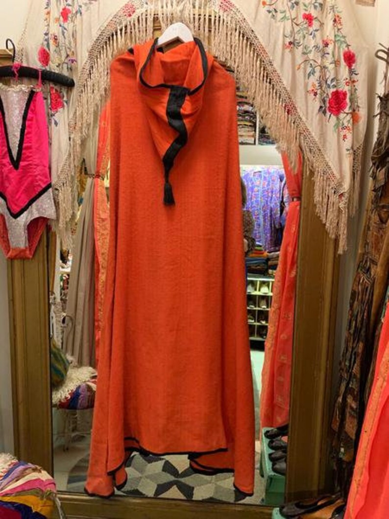 Puede incluir: Una capa con capucha naranja con ribete negro y borla. La capa est&aacute; colgada de una percha blanca en una tienda vintage. Un traje de ba&ntilde;o rosa con ribete negro cuelga a la izquierda.
