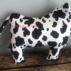 May include: Black and white cow-print plush toy with a long tail. The toy is standing on a wooden surface. The text "Rusty Needle Decor" is visible on the toy.