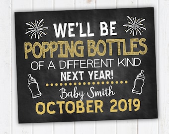 May include: Black chalkboard sign with gold text announcing a baby is due in October 2019. The text reads "We'll be popping bottles of a different kind next year! Baby Smith October 2019".