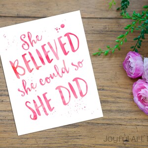 May include: A white print with the text "She BELIEVED she could so SHE DID" in pink watercolour lettering. The print is on a wooden surface, with pink flowers and greenery in the background.