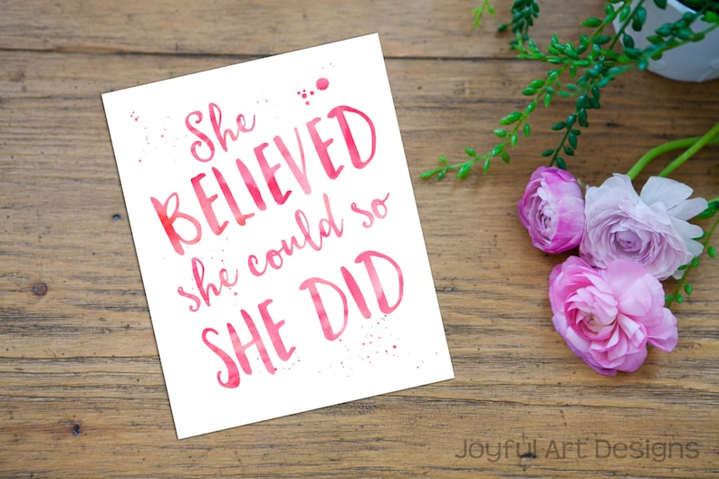 May include: A white print with the text "She BELIEVED she could so SHE DID" in pink watercolour lettering. The print is on a wooden surface, with pink flowers and greenery in the background.