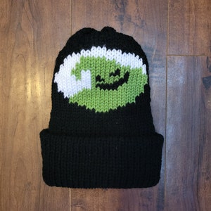 May include: Black knit beanie with a green and white knitted eye design. The eye has a white sclera and a green iris with a black pupil and a black smile.