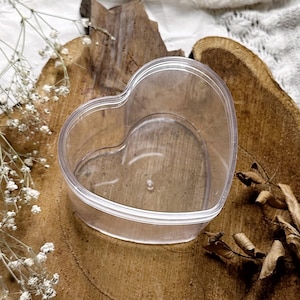 May include: A clear, heart-shaped plastic container sits on a rustic, circular wooden base. The container is empty, with a smooth, transparent surface. Dried leaves and delicate white flowers are arranged around the wooden base, creating a natural aesthetic.