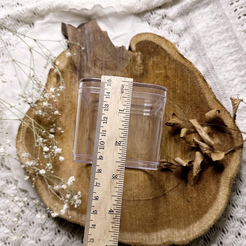 Op de afbeelding: Een heldere, rechthoekige plastic container wordt gemeten met een houten liniaal. De liniaal toont aan dat de container ongeveer 10 cm hoog is. De container staat op een rustieke, houten schijf met gedroogde bladeren en witte bloemen.