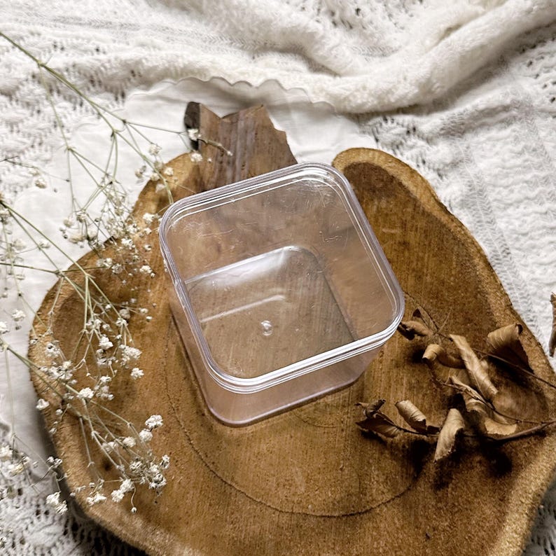 Op de afbeelding: Een doorzichtige, vierkante plastic container, geplaatst op een rustiek houten oppervlak. De container is leeg, waardoor de transparantie zichtbaar is. Gedroogde bladeren en kleine witte bloemen voegen een natuurlijk tintje toe.