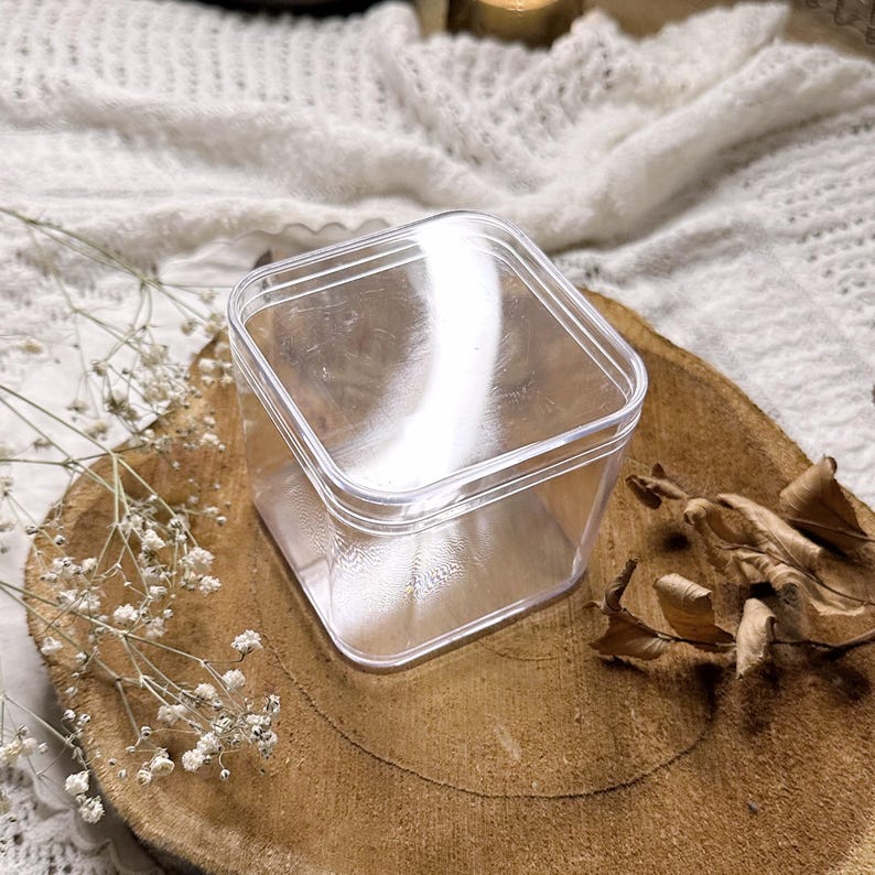 Op de afbeelding: Een doorzichtige, vierkante plastic container met een deksel. De container staat op een rond, houten oppervlak. Gedroogde bladeren en kleine witte bloemen zijn rond de container verspreid.
