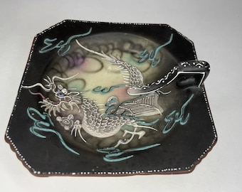 Könnte beinhalten: Eine schwarze Keramikschale mit einem silbernen Drachenmuster und einem Griff. Der Drache ist von blauen und weißen Wirbeln umgeben. Die Schale hat einen gewellten Rand.