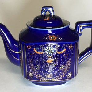 May include: A cobalt blue teapot with gold trim and a floral design. The teapot has a square body with a rounded lid and a handle.
