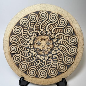 May include: A circular ceramic wall plaque featuring a central sun face with stylized facial features, surrounded by a radiating pattern of raised, dark brown clay spirals against a speckled, light beige, textured background surface.