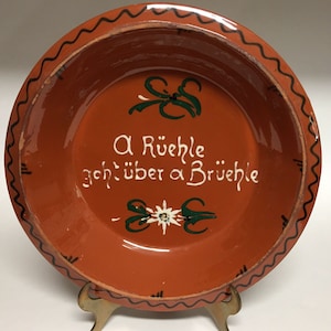 May include: A brown ceramic plate with a white and green floral design and the text "A Rüehle geht über a Brüehle".