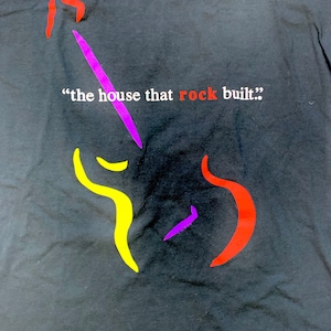 Op de afbeelding: Een zwart T-shirt met een abstract ontwerp in paars, geel en rood. De tekst "the house that rock built." is op het T-shirt gedrukt.