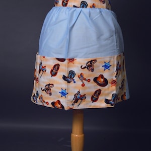 May include: A light blue apron with a white waistband and a pocket. The bottom half of the apron is a tan fabric with a pattern of cowboy boots, hats, and saddles.