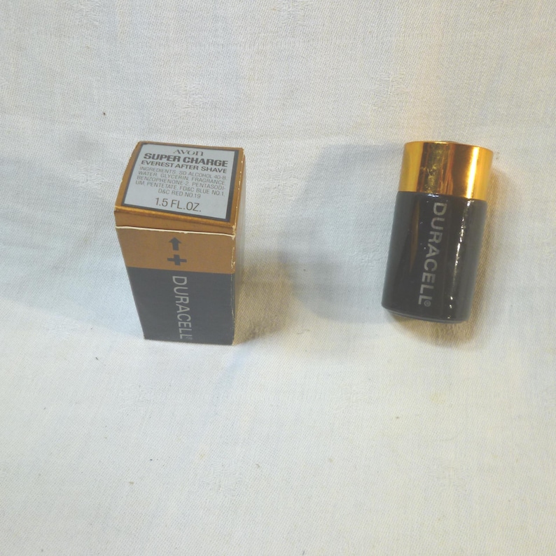 Op de afbeelding: Een vintage Avon Super Charge Everest aftershave fles en doos. De fles heeft de vorm van een Duracell-batterij, met een zwarte behuizing en een gouden dop. De doos is goud en zwart, met productinformatie en het Duracell-logo.