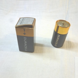 Op de afbeelding: Een Duracell D-cel batterij en de doos worden getoond op een wit oppervlak. De batterij is zwart met een gouden bovenkant en het Duracell logo. De doos is bruin en grijs met het Duracell logo en tekst.