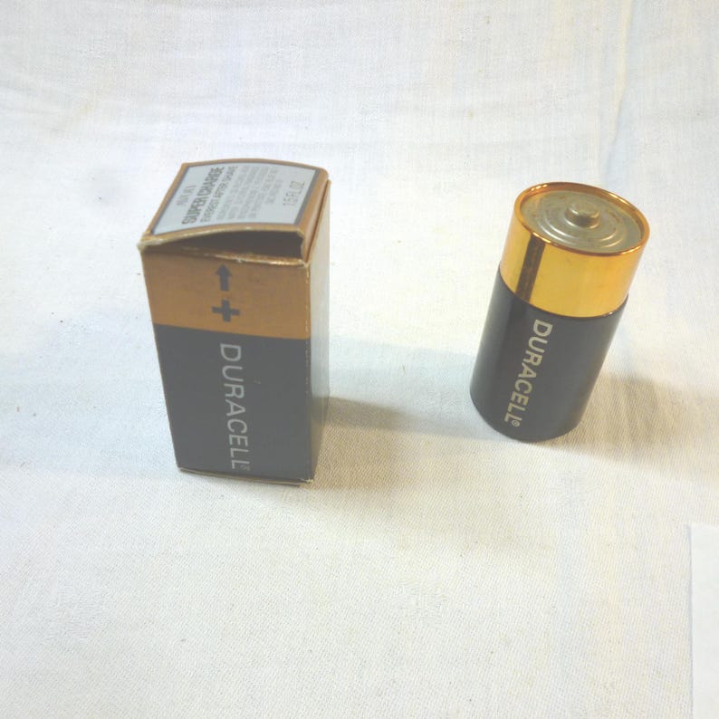 Op de afbeelding: Een Duracell D-cel batterij en de doos worden getoond op een wit oppervlak. De batterij is zwart met een gouden bovenkant en het Duracell logo. De doos is bruin en grijs met het Duracell logo en tekst.