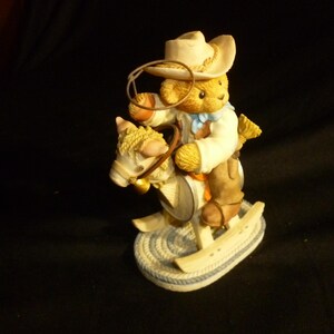 May include: A figurine of a teddy bear dressed as a cowboy, riding a rocking horse. The bear wears a cowboy hat, a white shirt, and brown chaps. The rocking horse is cream-colored with a blue and white base.