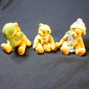 May include: Three small, seated teddy bear figurines. Each bear is a light brown color and wears a different accessory: a green hat and scarf, a bow, and a blue hat and scarf. The bears are on a dark background.