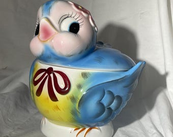 May include: A ceramic bird-shaped cookie jar with a removable lid. The bird is painted in shades of blue and yellow with a pink beak, large eyes with long eyelashes, and a red bow painted on the chest. It features a white base and a small flower.