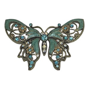 Puede incluir: Un broche de mariposa verde y dorado con diamantes de imitación transparentes y azules. La mariposa tiene un diseño de filigrana y está adornada con pequeñas flores doradas.