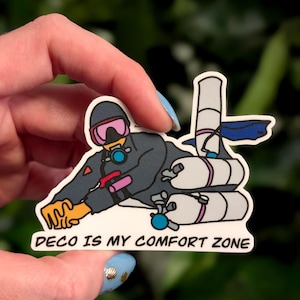 Puede incluir: Una pegatina con un buzo de dibujos animados con un traje de buceo gris oscuro, máscara rosa y guantes amarillos. El buzo está rodeado de tanques blancos y el texto "DECO IS MY COMFORT ZONE" en letras negras.