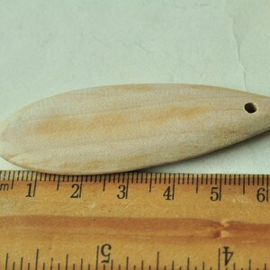 May include: A light brown wooden spoon with a hole at the top, measuring approximately 5 inches long. The spoon is resting on a wooden ruler.