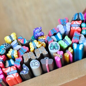 May include: A collection of colorful polymer clay butterfly canes in a wooden box. The canes feature various butterfly designs in shades of blue, pink, yellow, and green. The canes are cut into small pieces, showcasing the intricate patterns.