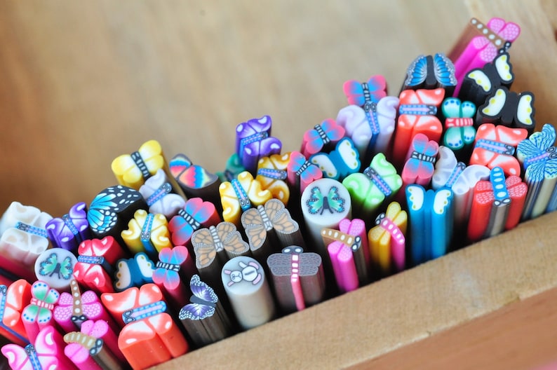 May include: A collection of colorful polymer clay butterfly canes in a wooden box. The canes feature various butterfly designs in shades of blue, pink, yellow, and green. The canes are cut into small pieces, showcasing the intricate patterns.