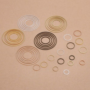 Op de afbeelding: Assortiment van metalen sprongringen in verschillende maten en kleuren, waaronder goud, zilver, brons en koper. De sprongringen zijn in een cirkelvormige opstelling op een bruine achtergrond geplaatst.