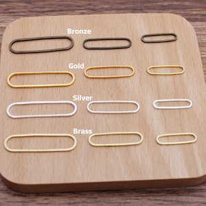 Puede incluir: Surtido de anillas ovaladas en cuatro acabados metálicos diferentes: bronce, oro, plata y latón. Las anillas están dispuestas en filas sobre una superficie de madera.