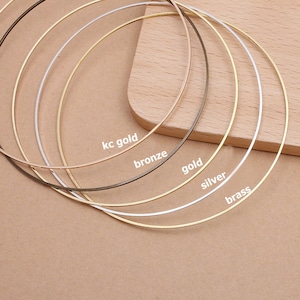 Peut inclure: Plusieurs colliers ras du cou en métal de différentes couleurs, dont or, argent, bronze et laiton. Les couleurs sont étiquetées : "kc gold", "bronze", "gold", "silver" et "brass". Les colliers sont disposés sur une surface marron clair.