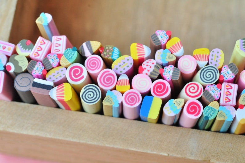 May include: A collection of colorful polymer clay canes, each intricately designed to resemble miniature desserts. The assortment includes cupcakes, ice cream cones, cakes, and spiral candies, all arranged in a wooden box. These are perfect for crafting.