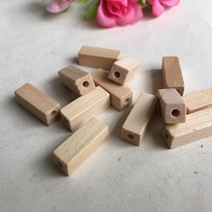 May include: A selection of unfinished wooden beads, rectangular with a central hole. The beads are a light, natural wood colour and are scattered on a neutral surface. Some beads have a slightly tapered end, and are ready for crafting.