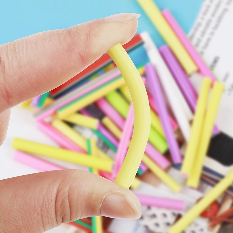 May include: A hand holding a curved, yellow, plastic straw. The background features a pile of colorful straws in various shades of yellow, pink, green, and purple. The straws appear to be for crafts or DIY projects.