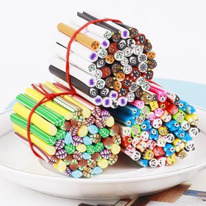 May include: Three bundles of colorful polymer clay canes, tied with red twine, rest on a white plate. The canes feature various designs, including leaves, Halloween-themed faces, and animal faces. The canes are approximately 6 inches long.