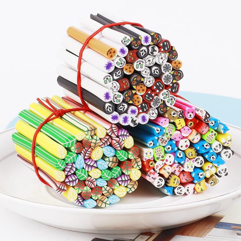 May include: Three bundles of colorful polymer clay canes, tied with red twine, rest on a white plate. The canes feature various designs, including leaves, Halloween-themed faces, and animal faces. The canes are approximately 6 inches long.
