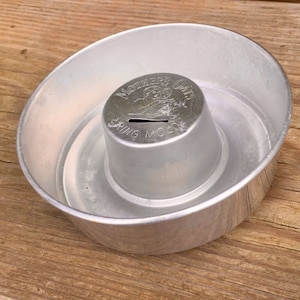 May include: A silver metal Mother's Oats cereal box shaped piggy bank with a slot on the top. The piggy bank is sitting on a wooden surface.