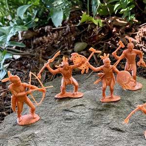 May include: A collection of orange plastic toy Native American figures, posed on a rock. The figures are in various action poses, some holding bows and arrows, others with shields and weapons. The figures are set against a backdrop of green foliage.
