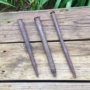 May include: Three antique metal chisels with a weathered, rusty appearance. The chisels are of varying shapes and sizes, with a tapered, pointed, or flat end. They are arranged on a weathered wooden surface, suggesting they are vintage tools.