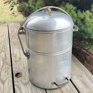 May include: A silver, two-tiered camping coffee pot with a handle and a lid. The pot has markings for measuring liquid.