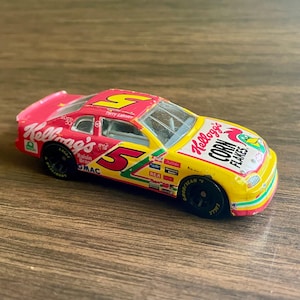 May include: A toy race car with a yellow and pink Kellogg's Corn Flakes design. The car has the number 5 on the side and the words "Terry Labonte" on the roof. The car has black wheels and a detailed exterior.