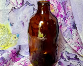 May include: A brown glass bottle with a rounded bottom and a slightly flared neck. The bottle is sitting on a purple and white floral fabric.