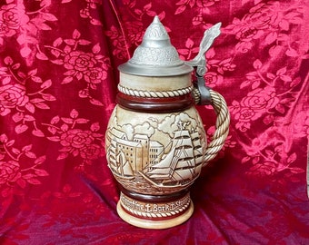 May include: A ceramic beer stein with a pewter lid and handle. The stein features a detailed scene of a ship sailing into a harbor. The text "Seefahrer T. Bark T. Stover" is inscribed on the stein.